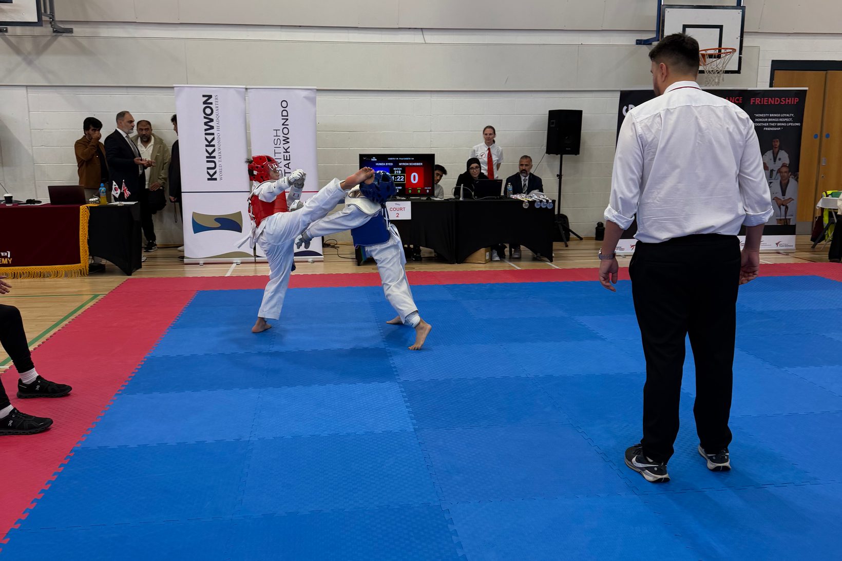 A fighter in a red chest guard throws a head kick during a bout on court 2
