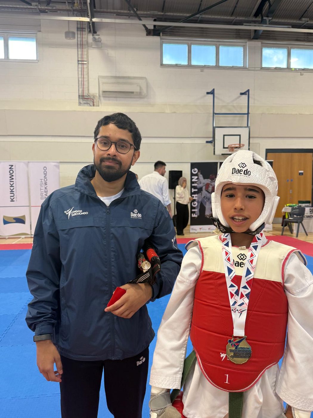 A young fighter wearing a gold medal after his bout, standing beside his coach
